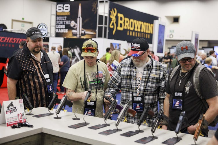 Men in caps examine pistols in a crowded convention.