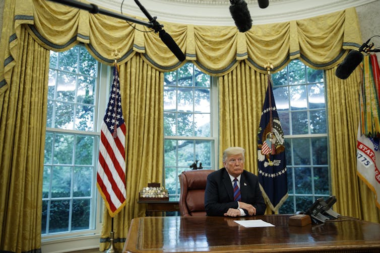 A man with sandy colored hair at a big desk in front of three large windows.