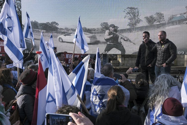 Several Israeli flags fly in front of a giant screen showing military action.