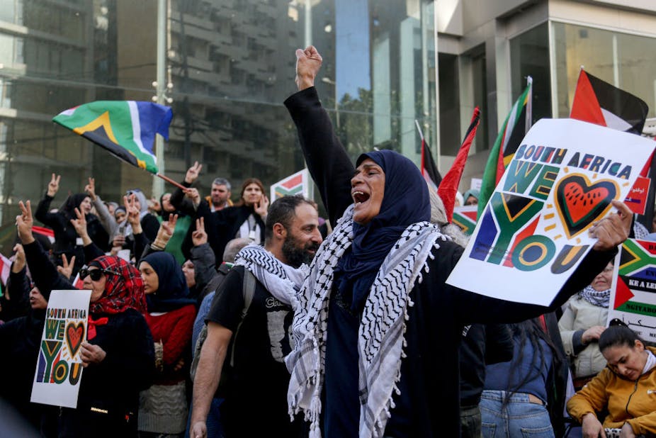 A group of protesters, many in Palestinian scarves, raise their hands and shout. In the centre, a woman holds a poster reading "South Africa we love you".