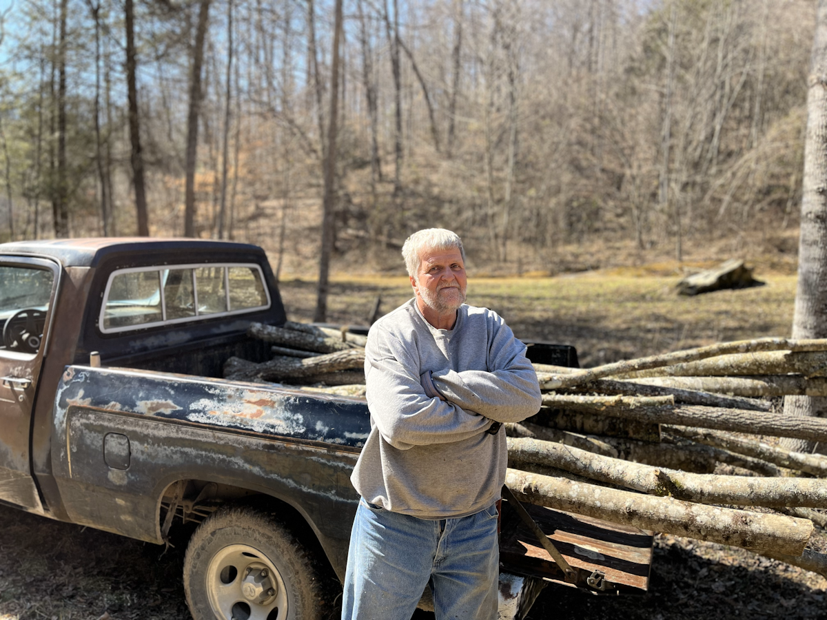 Randall Hatfield, a White man, stands in front of a pickup truck filled with cut timber.