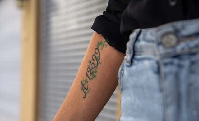 Una mujer vestida con vaqueros y camisa negra extiende el brazo en el que lleva tatuado un número.