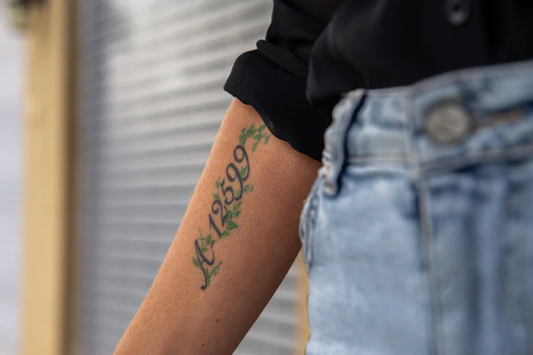 Una mujer vestida con vaqueros y camisa negra extiende el brazo en el que lleva tatuado un número.