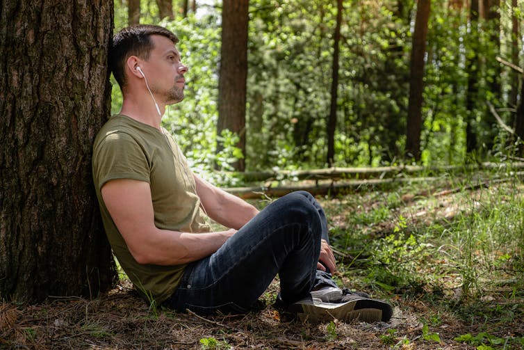 A man in tshirt and jeans sitting on the forest floor listening to earphones