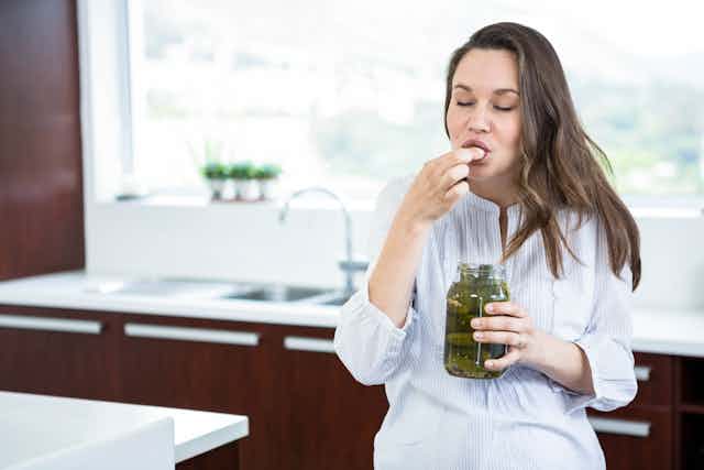 A pregnant woman eats a pickle from a jar.