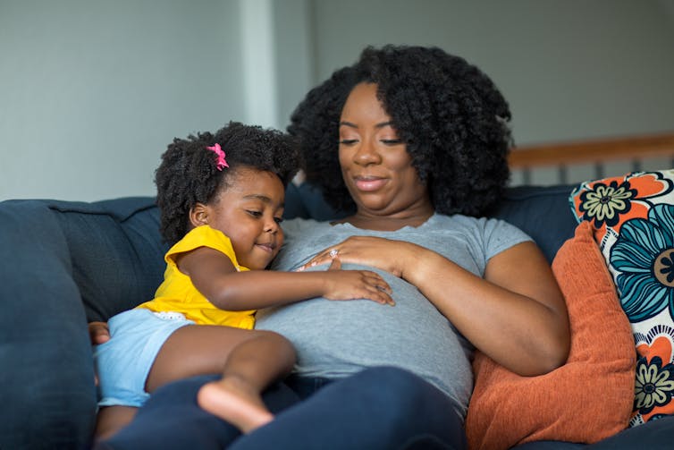 A pregnant woman on the couch with a toddler touching her stomach.