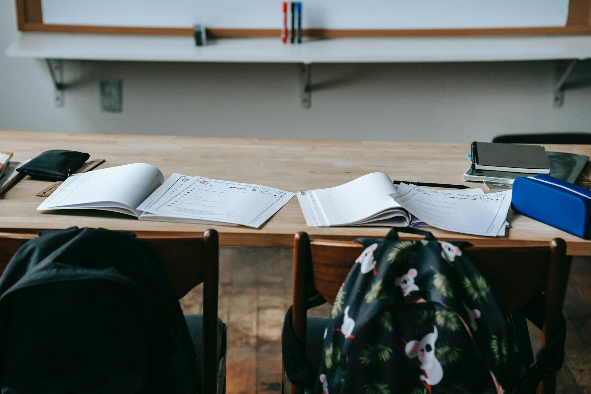 Backpacks on the back of chairs behind desks in a classroom. The desks have open books.