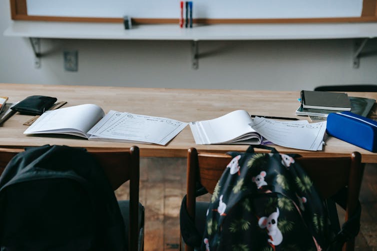 Backpacks on the back of chairs behind desks in a classroom. The desks have open books.