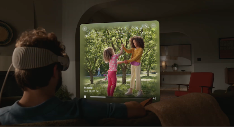 A man sitting on a couch and wearing a headset while a picture of children who play in the air swims in front of him.