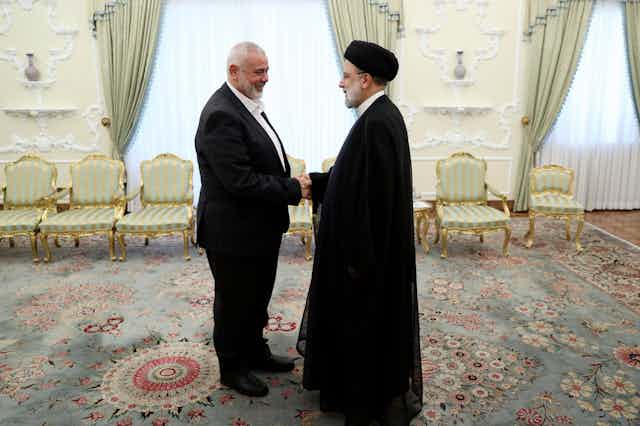 Two men shake hands in an ornate meeting room, one wearing a black turban and black robes..