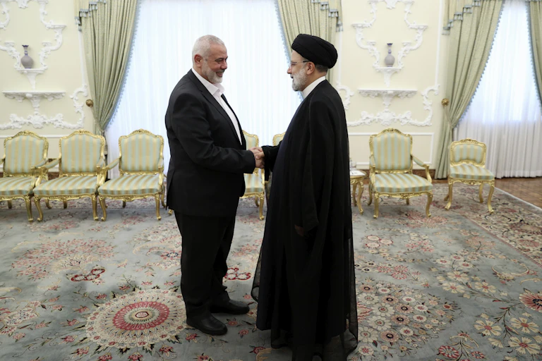 Two men shake hands in an ornate meeting room, one wearing a black turban and black robes..