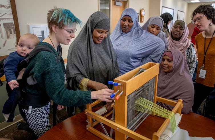 Several women in robes that cover their heads and bodies watch as a woman with a baby on her back demonstrates how to use a loom.