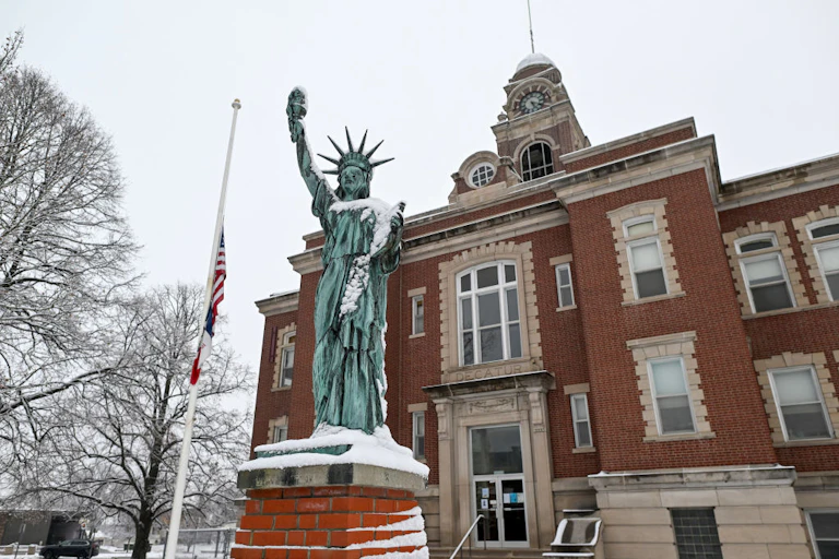 A green-gray statue is lightly covered in snow, standing in front of a brick building with a tower.