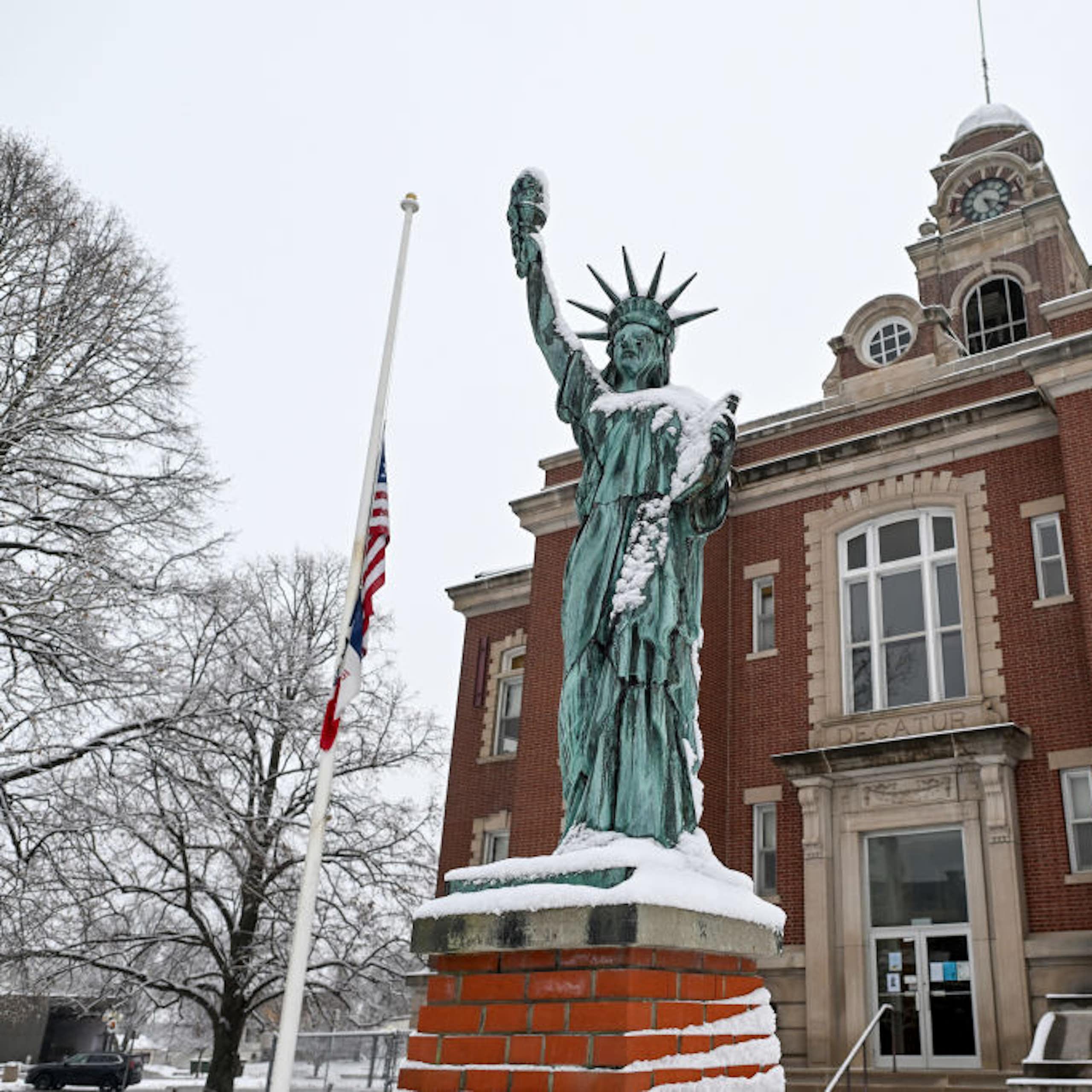 A green-gray statue is lightly covered in snow, standing in front of a brick building with a tower.