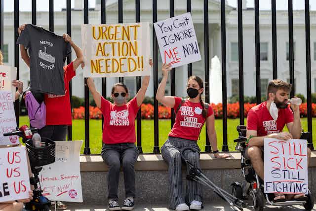 A group of people wearing red slogan t-shirts sit on a wall outside the White House holding banners in protest at the lack of investment in the medical condition ME/CFS