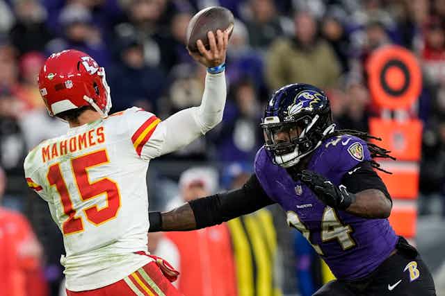 Quarterback for the Kansas City Chiefs holds a football while Baltimore Ravens linebacker presses him on defense.