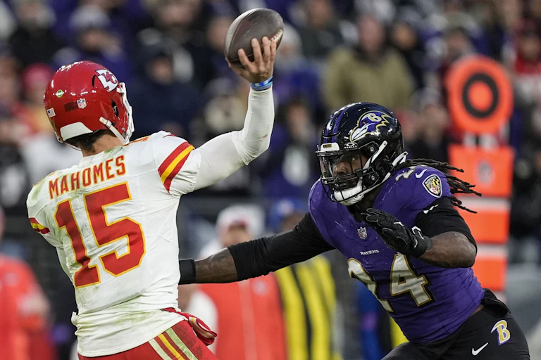 Quarterback for the Kansas City Chiefs holds a football while Baltimore Ravens linebacker presses him on defense.