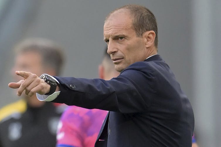 A middle-aged man with a receding hairline in a suit jacket points off-camera