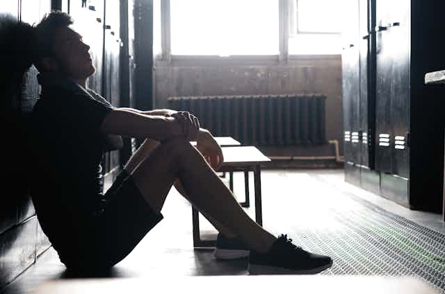 A young man sits with his back against a bank of lockers in a changing room