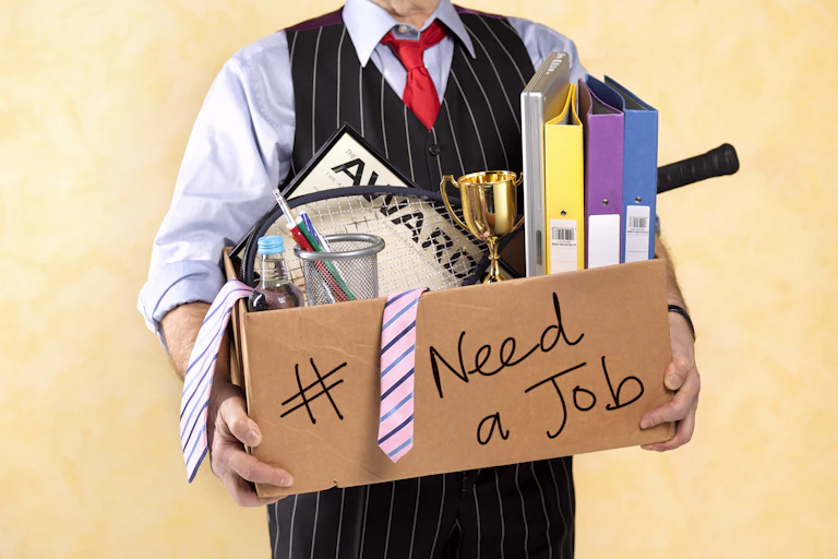 A businessman in a striped vest is seen holding a box that contains binders, ties and work awards. "Need a job" is scrawled on the box in marker.