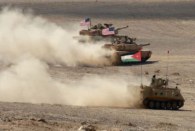 Tanks with Jordanian and US flags drive through the desert, kicking up dust.