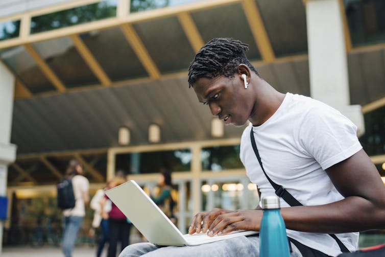 College student wearing ear buds works on laptop on campus