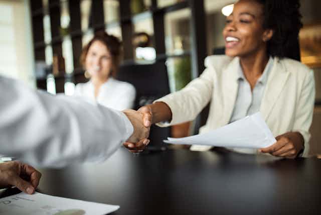 A businesswoman shakes hands across a boardroom table; her counterpart is mostly obscured.