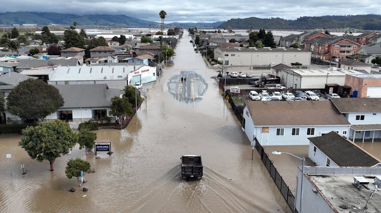 A truck drives through muddy streets that fill a large section of town. People stand on one small patch of pavement not flooded.