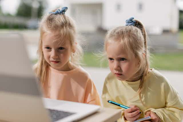 Young twin girls sit side by side, looking at a laptop. One holds a pen and pad.