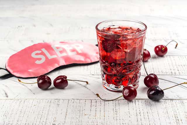 glass of red clear drink with sleep mask and cherries nearby on table
