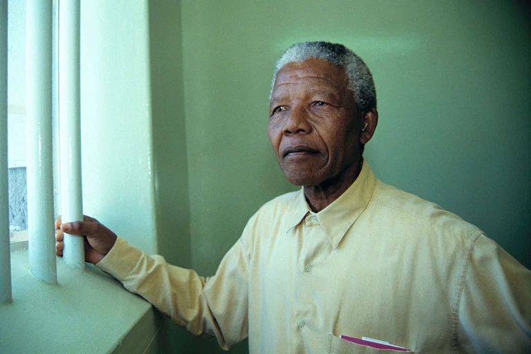 An elderly man stands beside a window, holding one of the window bars