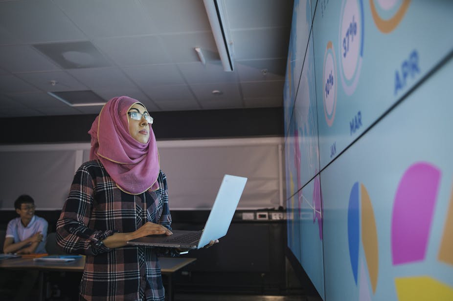 A young woman who is wearing a hijab uses a laptop to program an information wall for a presentation with colleagues.
