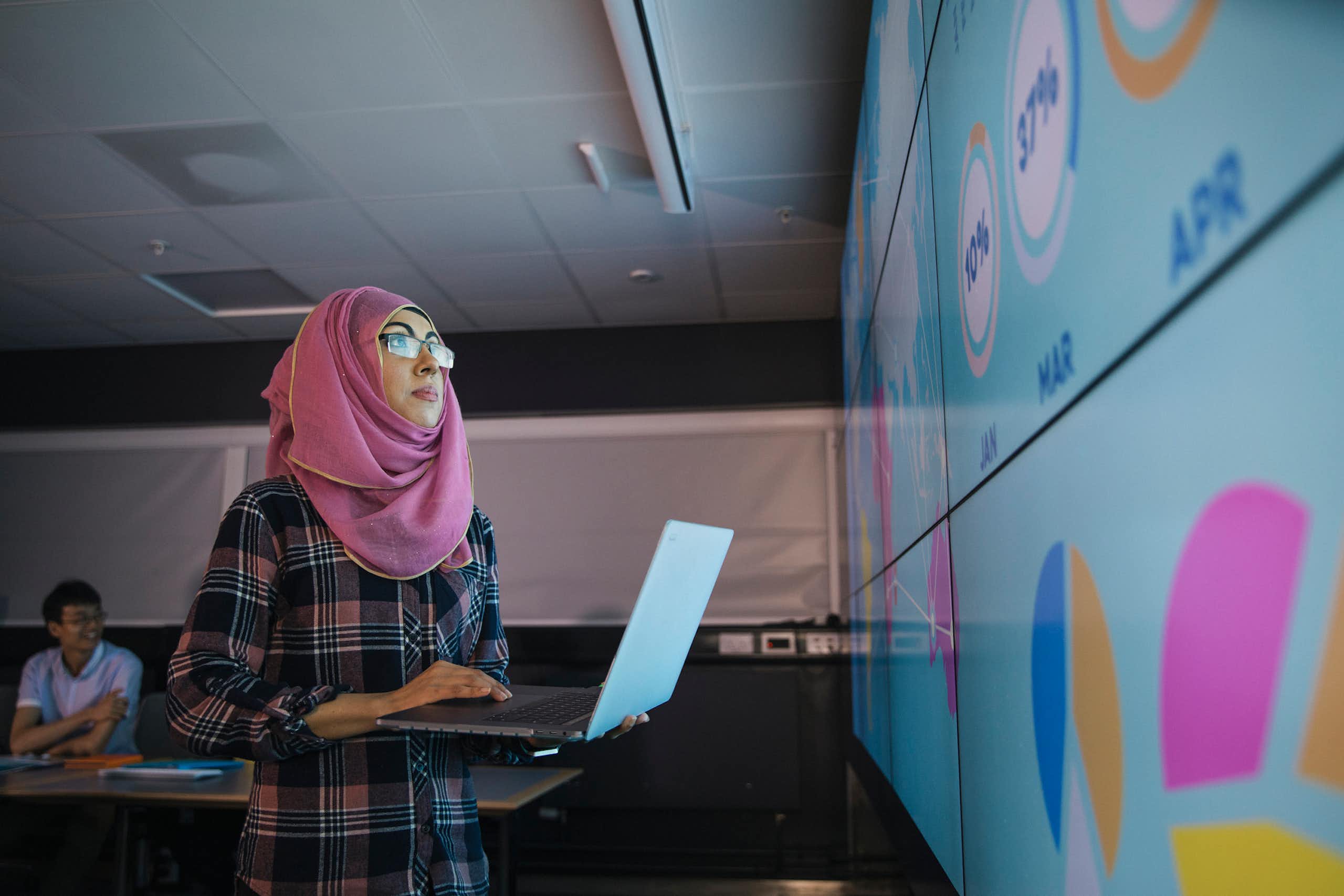 A young woman who is wearing a hijab uses a laptop to program an information wall for a presentation with colleagues.