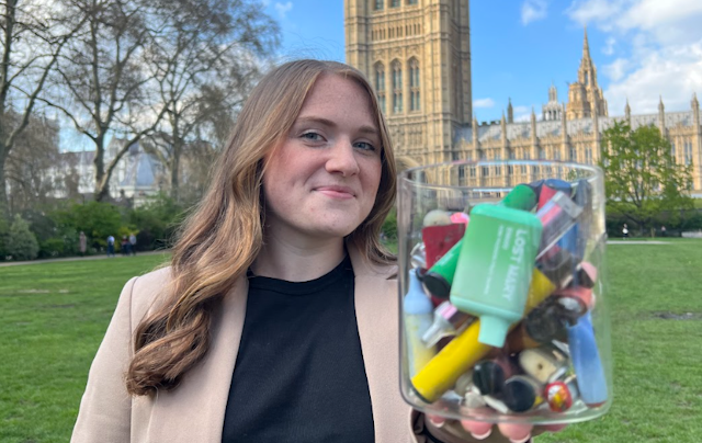 Woman with long brown hair holds a clear jar full of colourful littered vapes, against a background of Westminster with grand buildings and green lawn