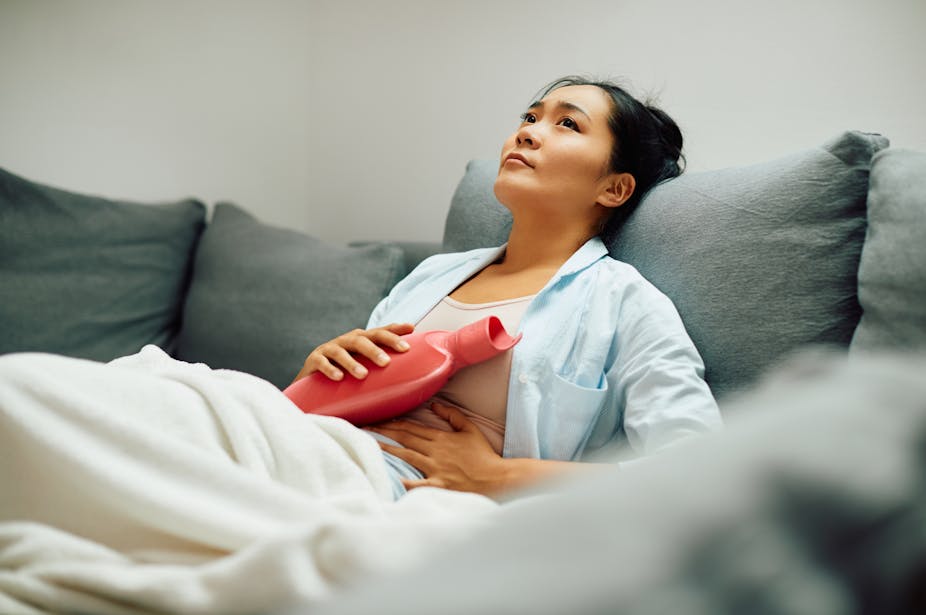 A woman sitting on a couch holds a hot water bottle on her abdomen.