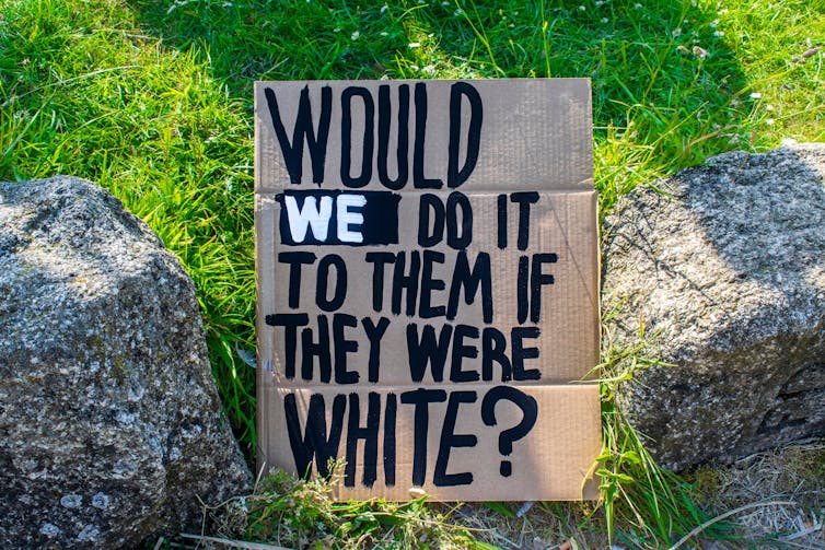 A cardboard sign sitting on the ground that reads 'would we do it to them if they were white?'