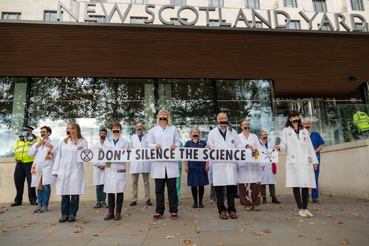 Scientists in white coats hold a sign saying 'don't silence the science' outside New Scotland Yard in London, UK.