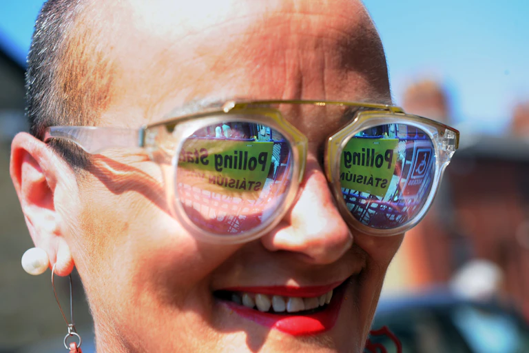 A woman outside a polling station with the "polling station" sign reflecting off her glasses.
