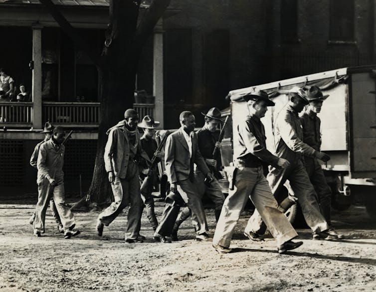 Several white men dressed in uniforms and carrying shotguns walk in front of a group of Black men.
