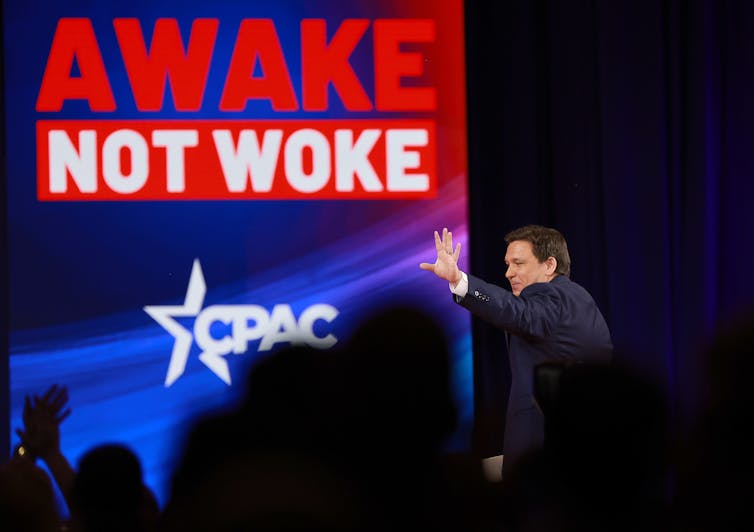 A white man waves to a crowd from a stage that has the words awake and not woke in large letters in the background.