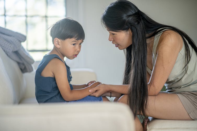 A woman with long black hair in shorts and a tank top sits as she talks and holds the hands of a young boy who also wears a tank top and shorts.