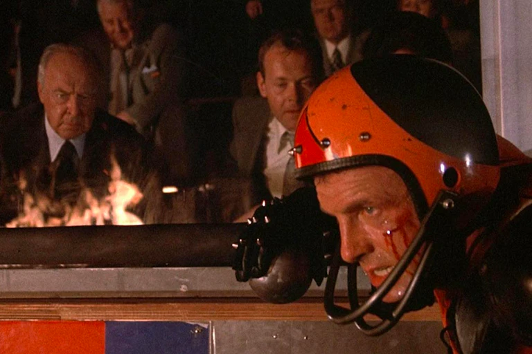 Movie still of two older men glaring at a bloodied athlete wearing a helmet.