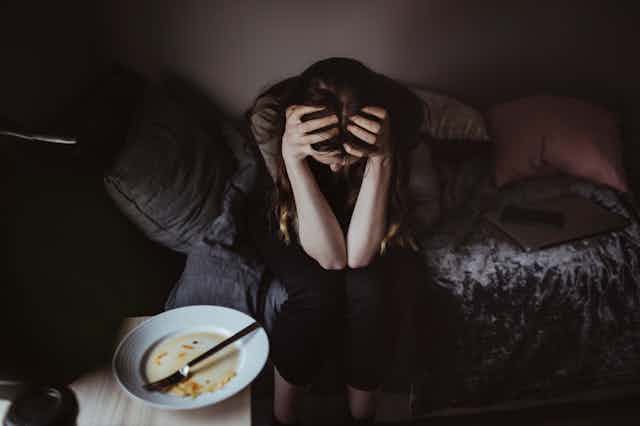 Person sitting on bed with elbows on knees and hands buried in their hair, an empty plate on the nightstand