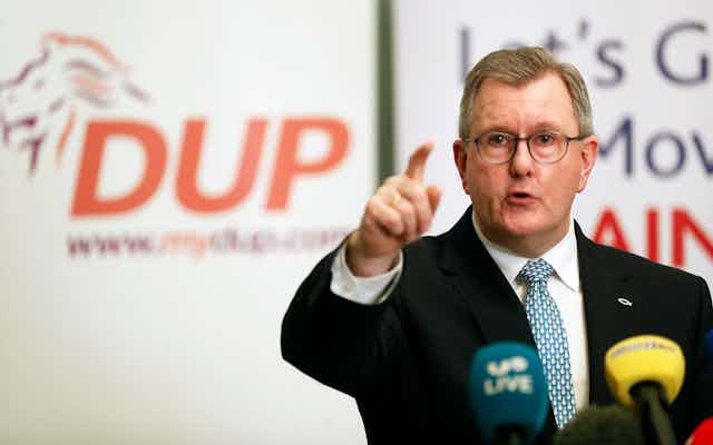 Jeffrey Donaldson pointing and speaking at a podium during a press conference, with the DUP logo on the wall behind him