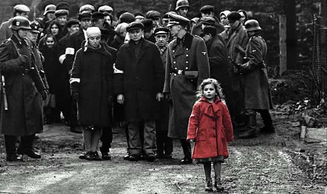 Fotografía en blanco y negro de prisioneros judíos con una niña con un abrigo rojo delante.