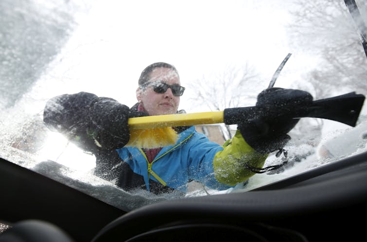 A person wearing a winter jacket uses a scraper on their frost-covered windshield.
