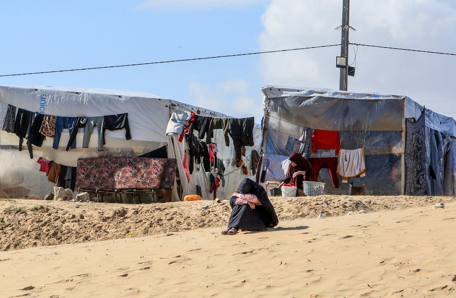 A woman sits on the ground with her head on her knees, with a makeshift tent and clothes drying on a line in the background.