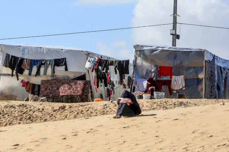 A woman sits on the ground with her head on her knees, with a makeshift tent and clothes drying on a line in the background.
