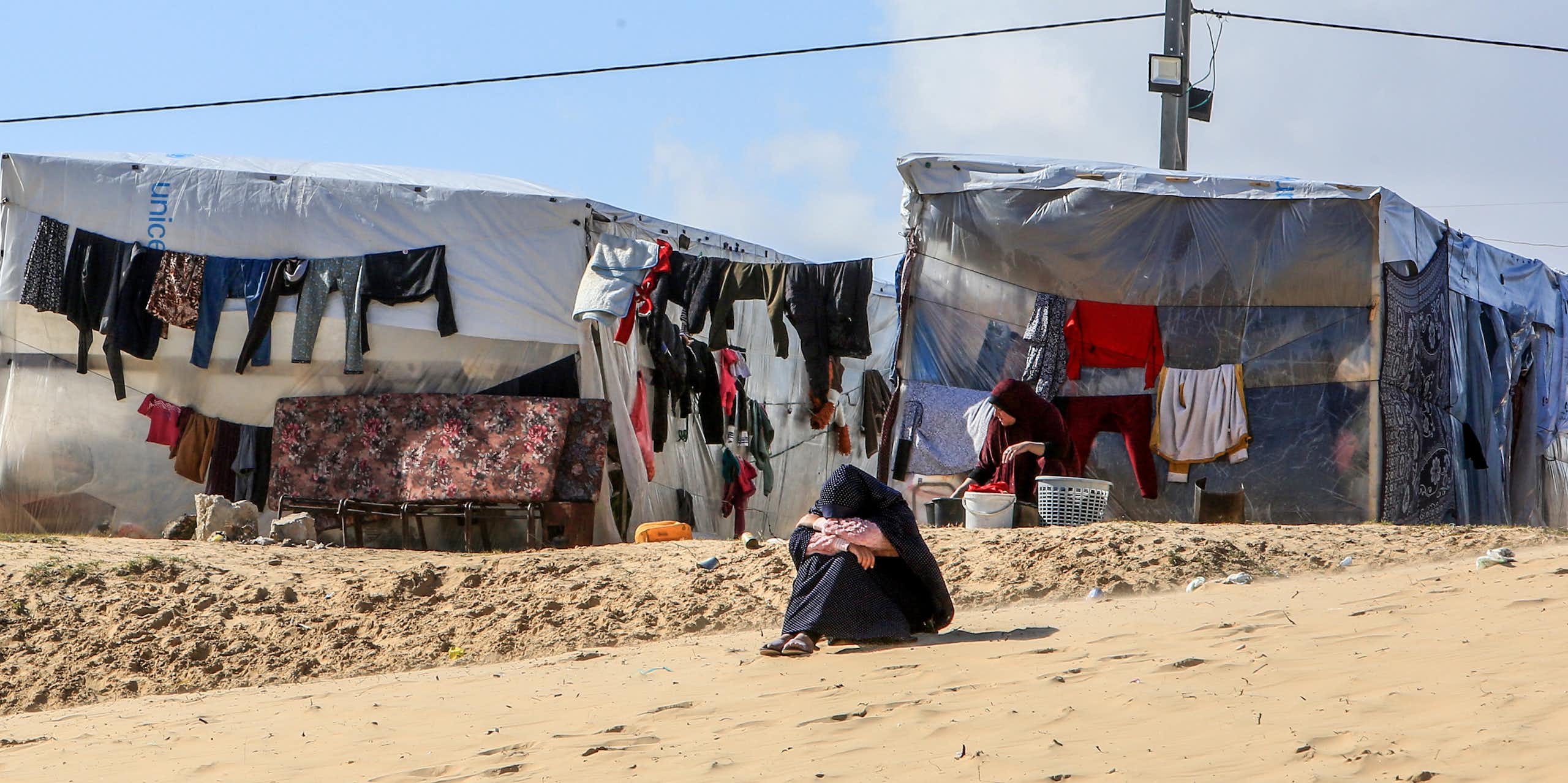 A woman sits on the ground with her head on her knees, with a makeshift tent and clothes drying on a line in the background.
