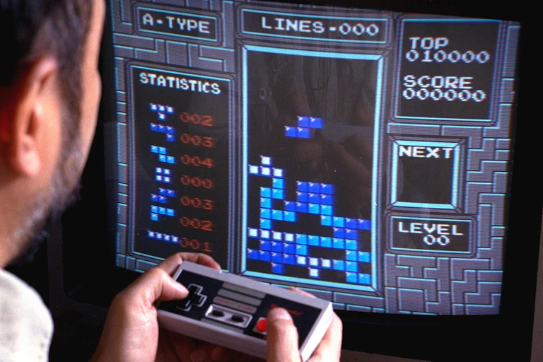 A man sitting in front of a TV screen displaying a 'Tetris' board with shapes stacking on top of each other. The man is holding a video game controller.